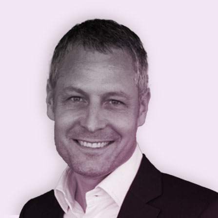 Smiling man with short gray hair wearing a suit jacket and white shirt, shown against a light purple background.