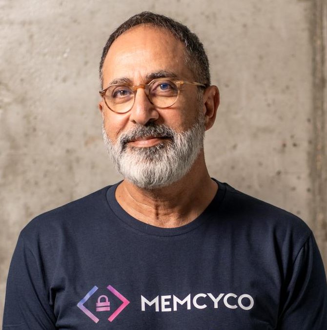 A man with glasses and a gray beard wears a navy blue MEMCYCO t-shirt, standing in front of a concrete wall, representing Memcyco at RSA Conference 2025.